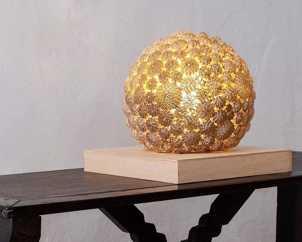 Escultura de luz con forma de esfera elaborada con cartón ondulado, proyectando luz cálida a través de patrones concéntricos.